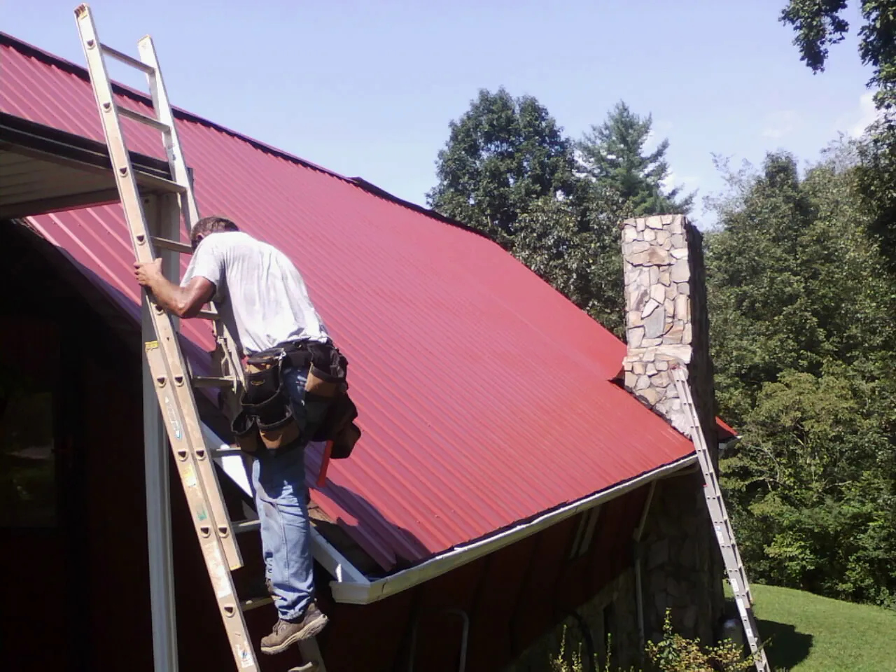 Roofer on a ladder beside a red metal roof and stone chimney — Soffit & Fascia Repair by Roofing Brothers in Walsenburg